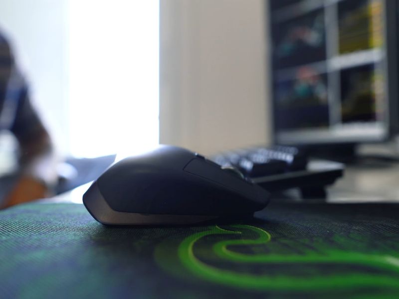 Bežični miš stoji na zeleno sivoj podlozi za miš, u pozadini tastatura i monitor