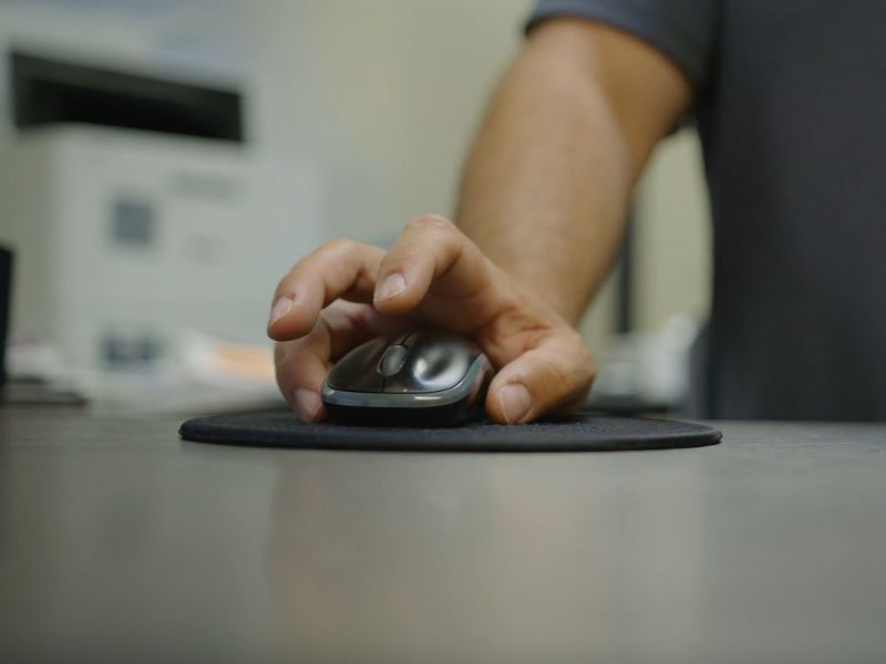 Osoba koristi miš za komjuter, miš stoji na crnoj podlozi koja se nalazi na sivom stolu